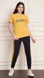 A woman smiling and standing against a white paneled wall. She is wearing a yellow Lycra cotton t-shirt with the word "QUEEN" printed on the chest, and black track pants with a yellow stripe down the side, also printed with the word "QUEEN". She is wearing white sneakers.