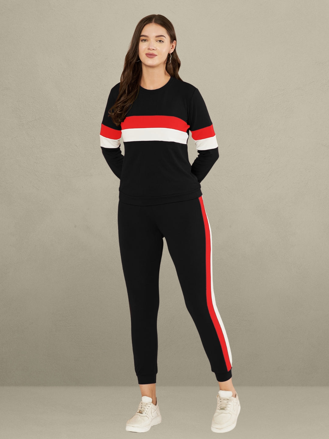 A woman standing against a plain gray background. She is wearing a red full-sleeve t-shirt with horizontal black and white stripes on the chest and sleeves, paired with matching red track pants that have a vertical black and white stripe down the side of each leg. She is also wearing white sneakers.