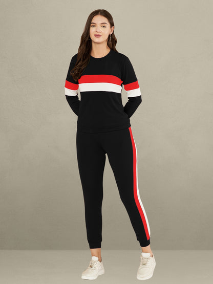 A woman standing against a plain gray background. She is wearing a red full-sleeve t-shirt with horizontal black and white stripes on the chest and sleeves, paired with matching red track pants that have a vertical black and white stripe down the side of each leg. She is also wearing white sneakers.