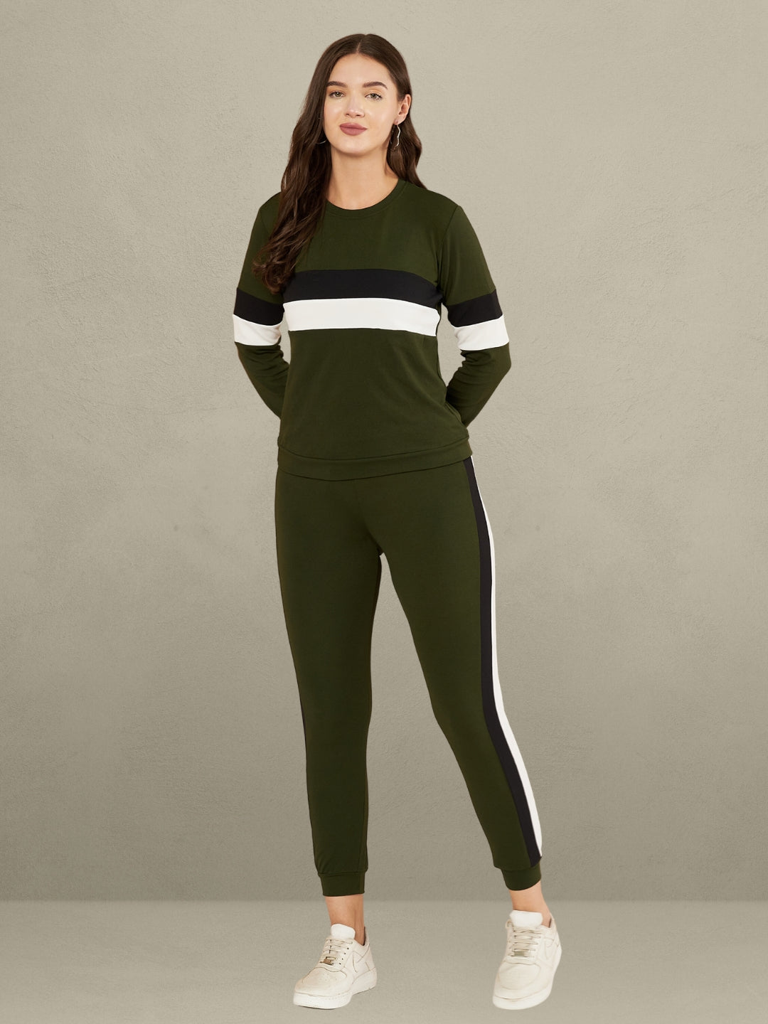 A woman standing against a plain gray background. She is wearing a green full-sleeve t-shirt with horizontal black and white stripes on the chest and sleeves, paired with matching green track pants that have a vertical black and white stripe down the side of each leg. She is also wearing white sneakers.