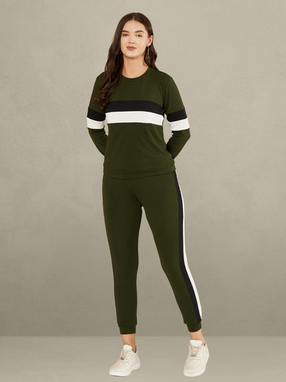 A woman standing against a plain gray background. She is wearing a green full-sleeve t-shirt with horizontal black and white stripes on the chest and sleeves, paired with matching green track pants that have a vertical black and white stripe down the side of each leg. She is also wearing white sneakers.