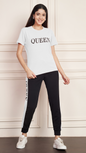 A woman smiling and standing against a white paneled wall. She is wearing a white Lycra cotton t-shirt with the word "QUEEN" printed on the chest, and black track pants with a white stripe down the side, also printed with the word "QUEEN". She is wearing white sneakers.