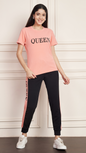 A woman smiling and standing against a white paneled wall. She is wearing a pink Lycra cotton t-shirt with the word "QUEEN" printed on the chest, and black track pants with a pink stripe down the side, also printed with the word "QUEEN". She is wearing white sneakers.