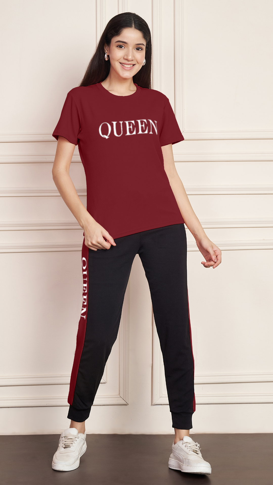 A woman smiling and standing against a white paneled wall. She is wearing a red Lycra cotton t-shirt with the word "QUEEN" printed on the chest, and blacktrack pants with a red stripe down the side, also printed with the word "QUEEN". She is wearing white sneakers.