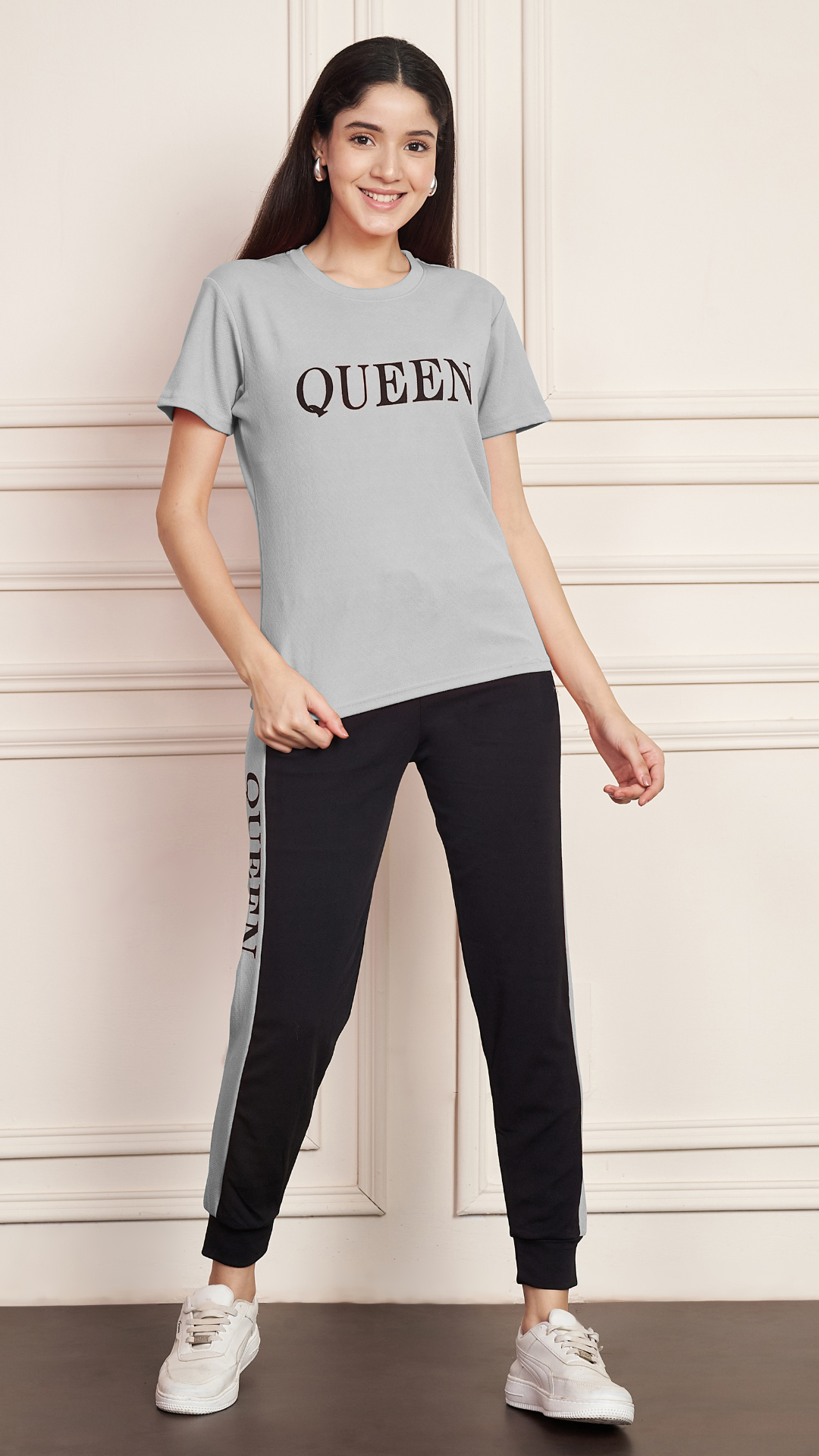 A woman smiling and standing against a white paneled wall. She is wearing a grey Lycra cotton t-shirt with the word "QUEEN" printed on the chest, and black track pants with a grey stripe down the side, also printed with the word "QUEEN". She is wearing white sneakers.