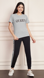 A woman smiling and standing against a white paneled wall. She is wearing a grey Lycra cotton t-shirt with the word "QUEEN" printed on the chest, and black track pants with a grey stripe down the side, also printed with the word "QUEEN". She is wearing white sneakers.