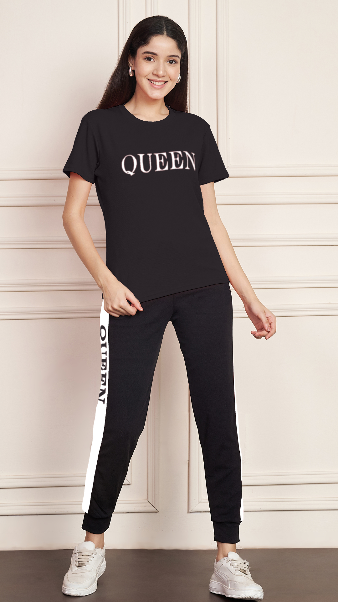 A woman smiling and standing against a white paneled wall. She is wearing a black Lycra cotton t-shirt with the word "QUEEN" printed on the chest, and black track pants with a white stripe down the side, also printed with the word "QUEEN". She is wearing white sneakers.