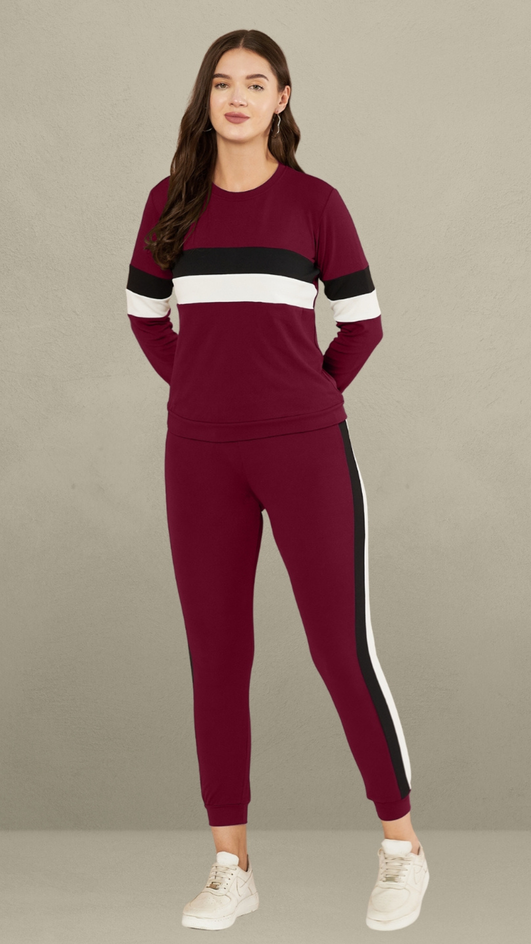A woman standing against a plain gray background. She is wearing a maroon full-sleeve t-shirt with horizontal black and white stripes on the chest and sleeves, paired with matching maroon track pants that have a vertical black and white stripe down the side of each leg. She is also wearing white sneakers.